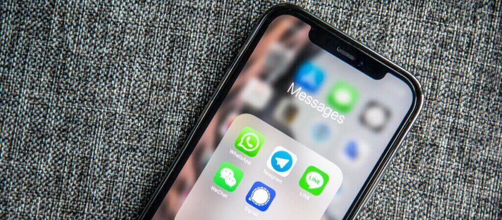 A smartphone lying on a gray fabric surface displaying a “Messages” folder with messaging apps including WhatsApp, Telegram, WeChat, Signal, and LINE.