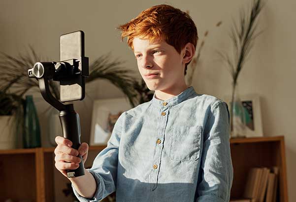 A young boy with reddish-brown hair is holding a smartphone mounted on a selfie stick. He is looking slightly off-camera. The background is a softly lit room with shelves and plants. A young boy with reddish-brown hair is holding a smartphone mounted on a selfie stick. He is looking slightly off-camera. The background is a softly lit room with shelves and plants.