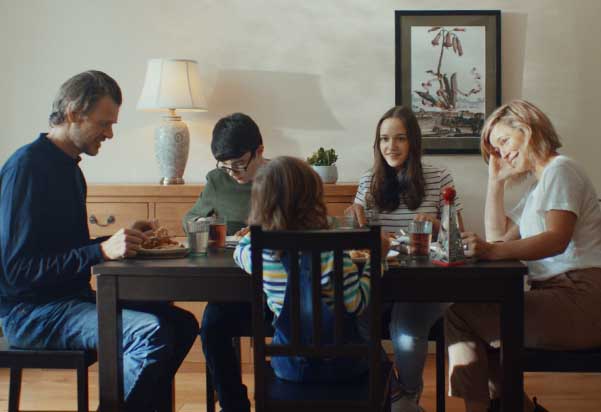 A family of five is seated around a dark wooden dining table, having a meal. They are engaged in conversation and eating, with plates and drinks on the table. In the background, there's a sideboard with a lamp and a framed picture on the wall. A family of five is seated around a dark wooden dining table, having a meal. They are engaged in conversation and eating, with plates and drinks on the table. In the background, there's a sideboard with a lamp and a framed picture on the wall.