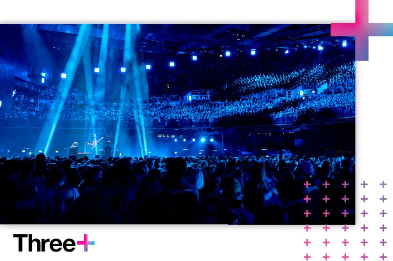 A crowded live concert at the 3Arena indoor venue bathed in bright blue stage lights, framed by a Three+ loyalty program branded border.