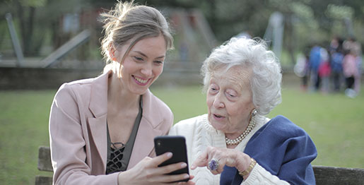 A smiling young woman with her hair pulled back is holding a black smartphone and showing it to an older woman. A smiling young woman with her hair pulled back is holding a black smartphone and showing it to an older woman.