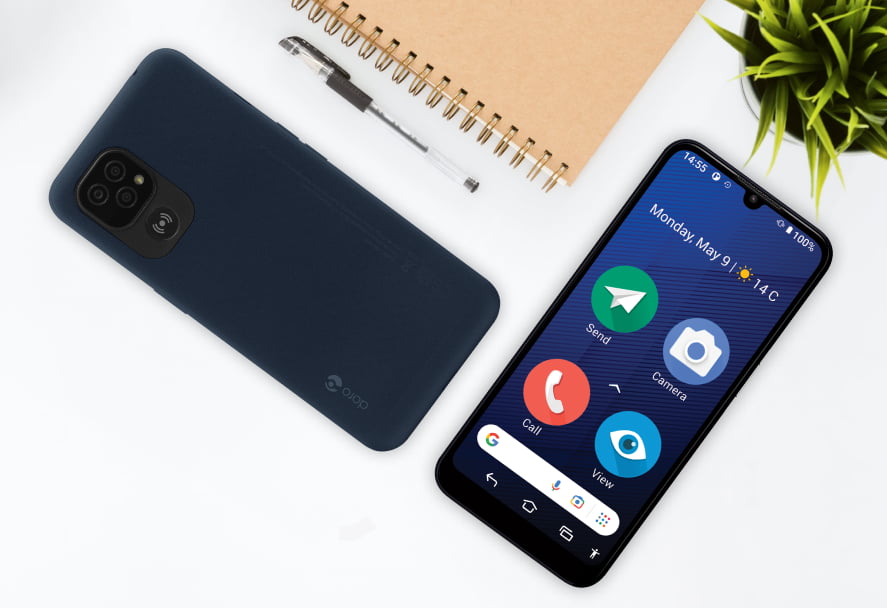 A dark blue smartphone, with its back cover and front screen visible, lying on a white surface. Beside the phone, there's a spiral-bound notebook, a pen, and a small green plant. A dark blue smartphone, with its back cover and front screen visible, lying on a white surface. Beside the phone, there's a spiral-bound notebook, a pen, and a small green plant.