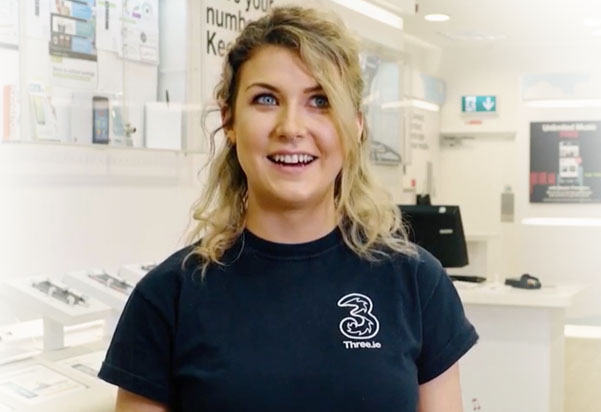 A blonde woman with blue eyes is smiling and looking slightly upward. She is wearing a dark blue t-shirt with a white "3" logo and "Three.ie" written underneath it. She appears to be in a retail store environment, with display shelves and a counter. A blonde woman with blue eyes is smiling and looking slightly upward. She is wearing a dark blue t-shirt with a white "3" logo and "Three.ie" written underneath it. She appears to be in a retail store environment, with display shelves and a counter.