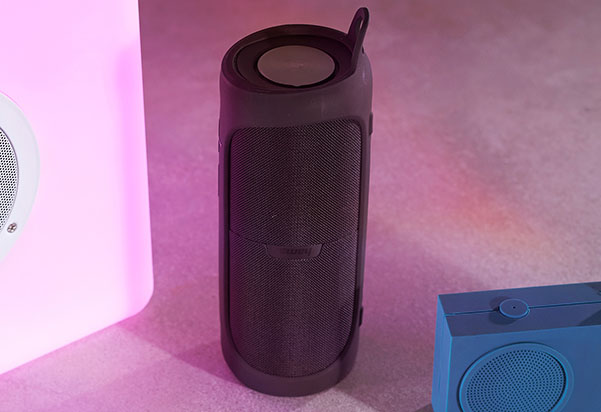 A dark charcoal portable Hama Bluetooth speaker standing upright with a carry loop on top, surrounded by colorful pink and blue speakers in a softly lit product display.
