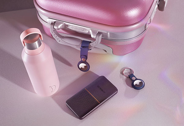 A styled flat lay of pink travel accessories including a hardshell carry-on suitcase, an insulated water bottle, a portable external hard drive, and two Apple AirTags, arranged on a light surface with soft iridescent reflections.