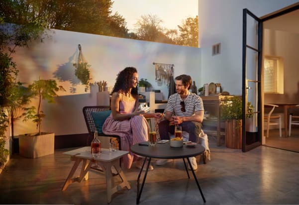 Two people relaxing on a sunlit outdoor patio, smiling and sharing a moment together, with a Polaroid Now instant camera nearby — capturing the ease of spontaneous, everyday photography.