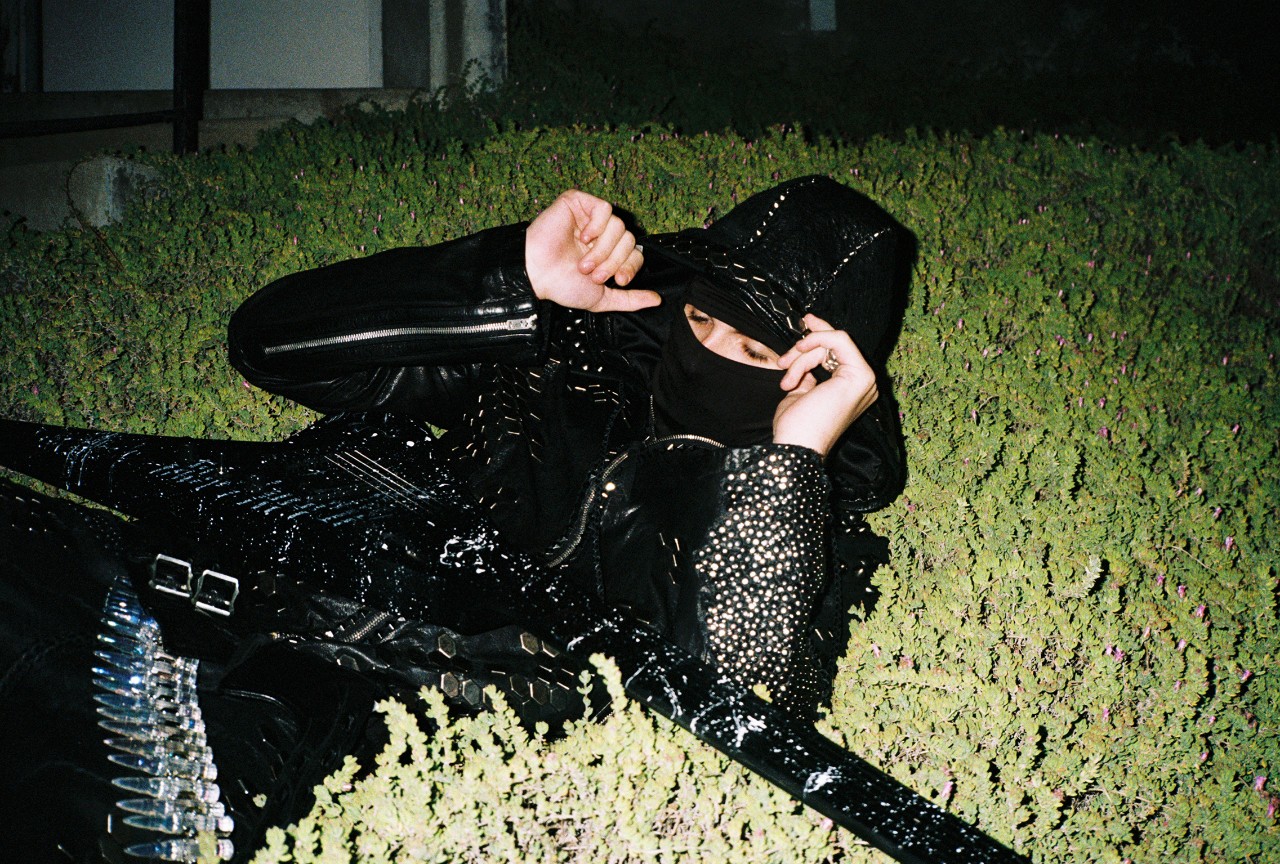 Esdeekid wearing a black hooded outfit with a face covering and thigh-high patent boots, posing among green hedges in a promotional image for their tour