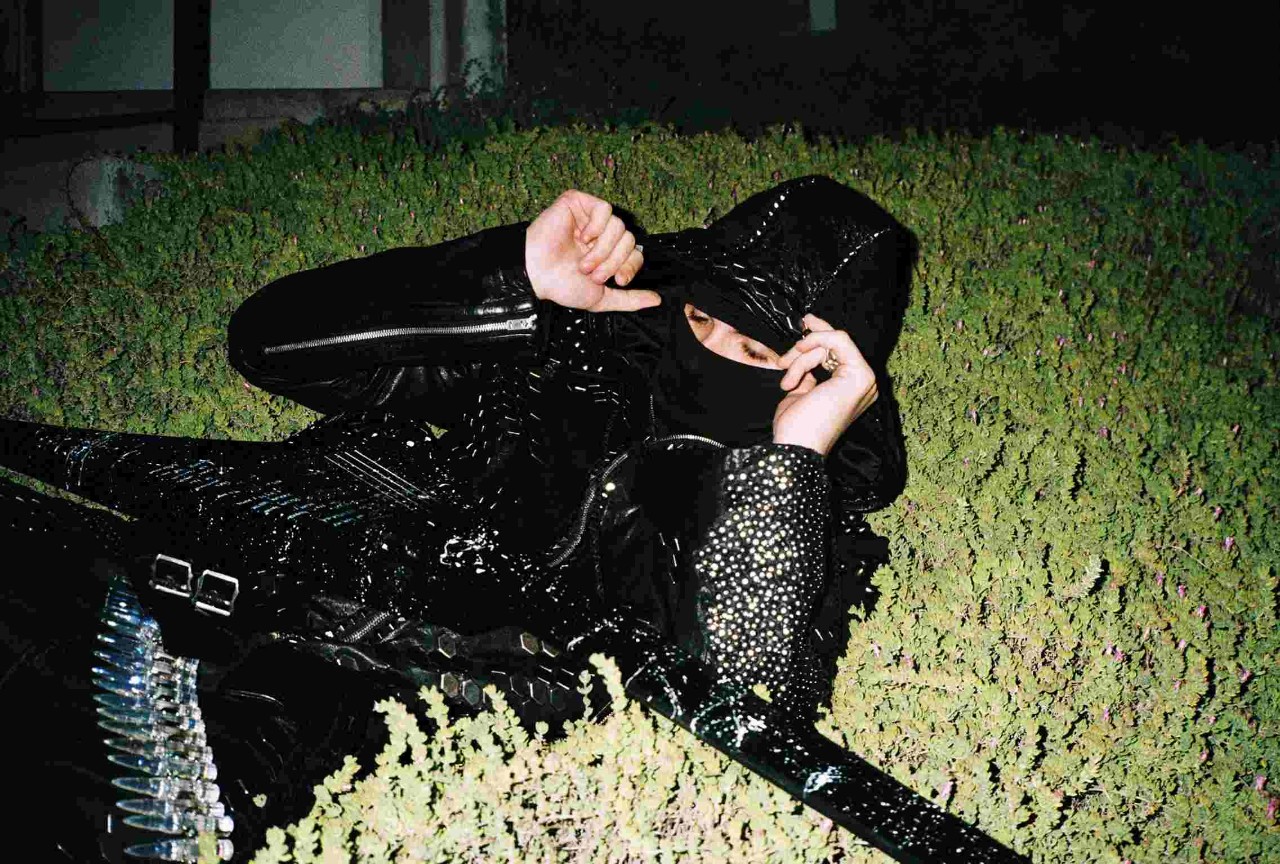 Esdeekid wearing a black hooded outfit with a face covering and thigh-high patent boots, posing among green hedges in a promotional image for their tour