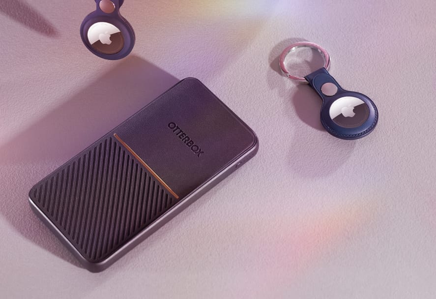 An Otterbox portable external hard drive in dark textured casing with a gold accent, laid flat on a soft surface alongside two Apple AirTags on keyrings, with soft iridescent light reflections.