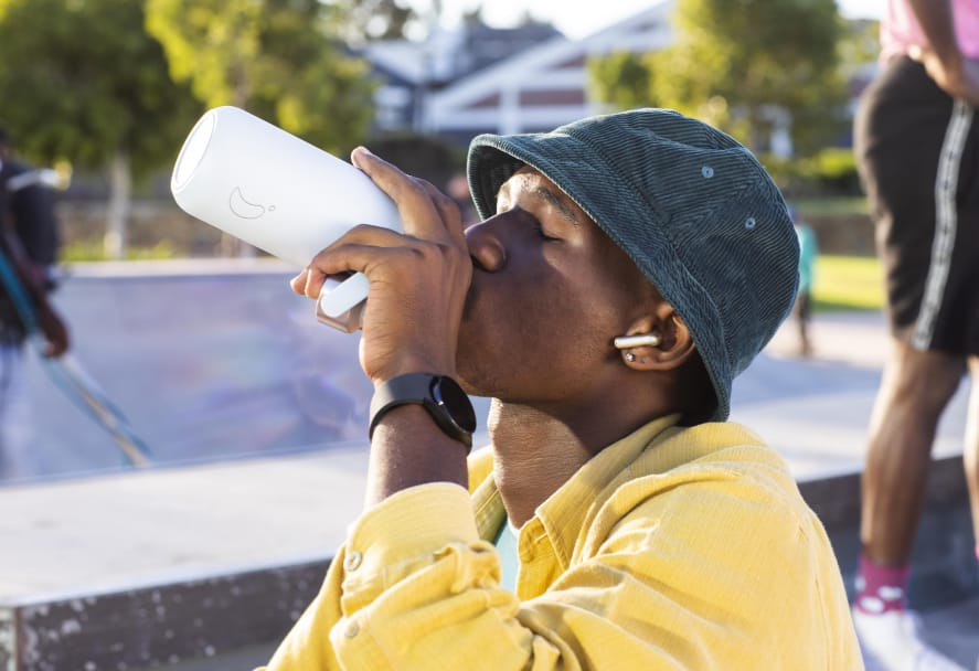 A young person in a yellow jacket and blue bucket hat drinking from a white insulated water bottle at an outdoor skate park on a sunny day, with wireless earbuds in and a smartwatch on their wrist.