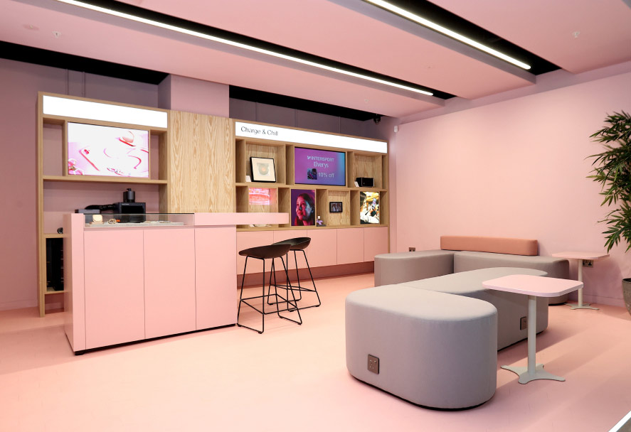 A contemporary space in the Three Dundrum store with shades of pink and light wood features a counter, shelves displaying items and screens, a charging station, and modular light grey seating with pink accents.