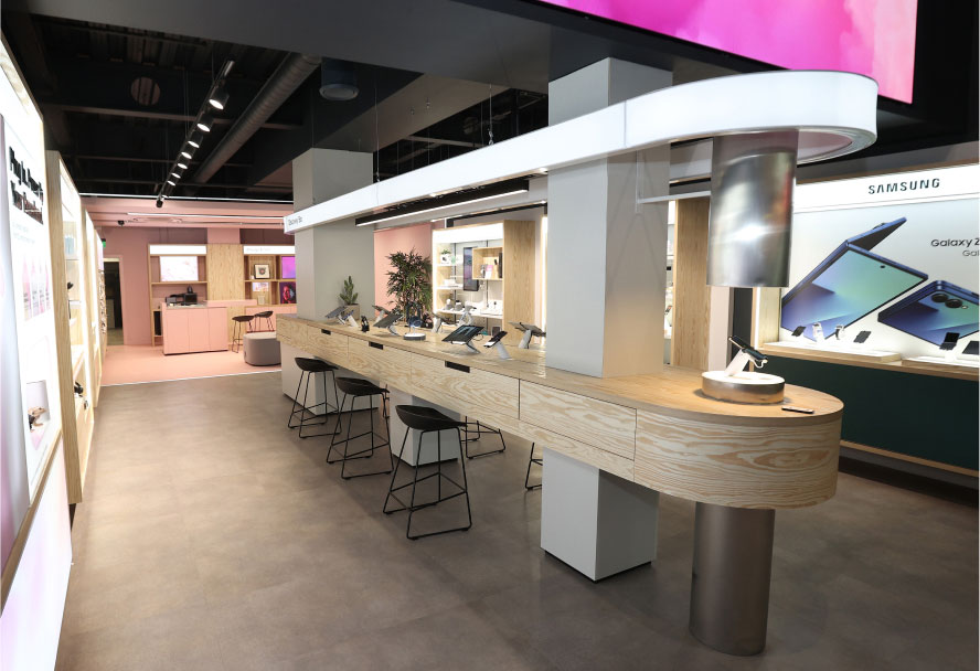 A bright, modern Three store interior featuring a long, light wood counter with stools and display phones, surrounded by display cases with devices, all under bright strip lighting.