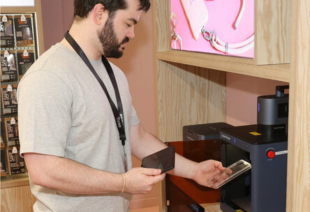A Three store employee, wearing a grey t-shirt and Three lanyard, stands in a store, holding a phone in one hand in a clear phone case and a tablet with the other.