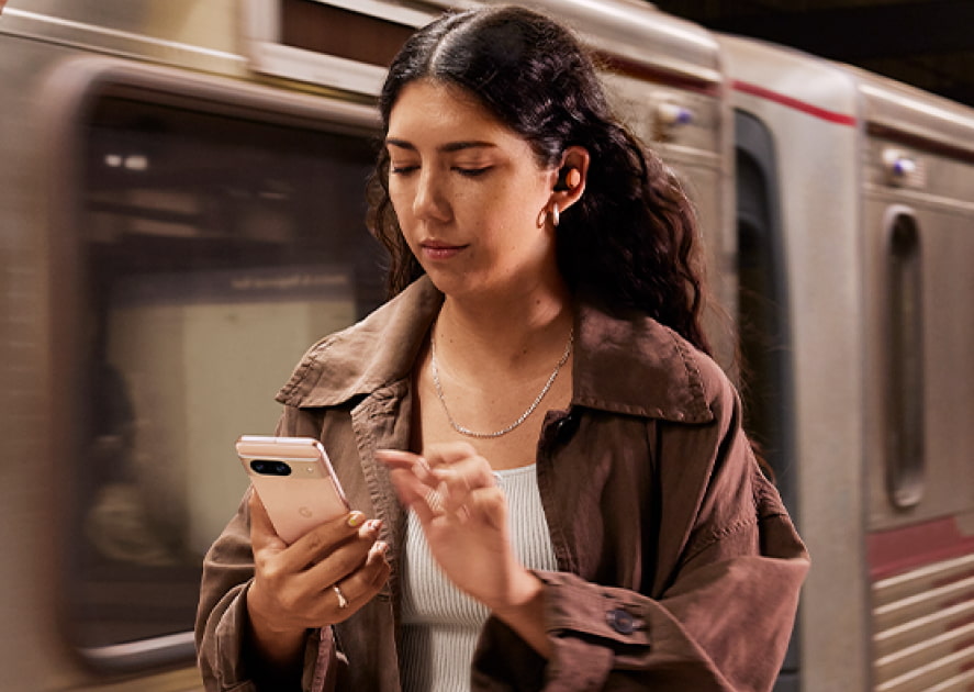 A young woman with long, dark wavy hair is looking down at a light-colored smartphone she is holding in her left hand, while her right hand is poised to touch the screen. In the background a train is visible. A young woman with long, dark wavy hair is looking down at a light-colored smartphone she is holding in her left hand, while her right hand is poised to touch the screen. In the background a train is visible.