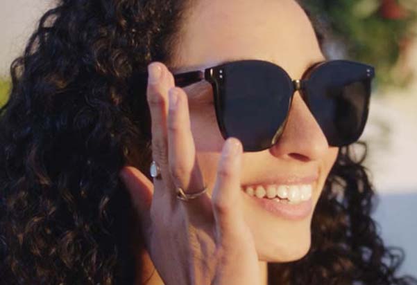 A smiling woman with curly hair wearing black Huawei smart sunglasses, touching the frame to interact with the built-in controls on a bright sunny day.