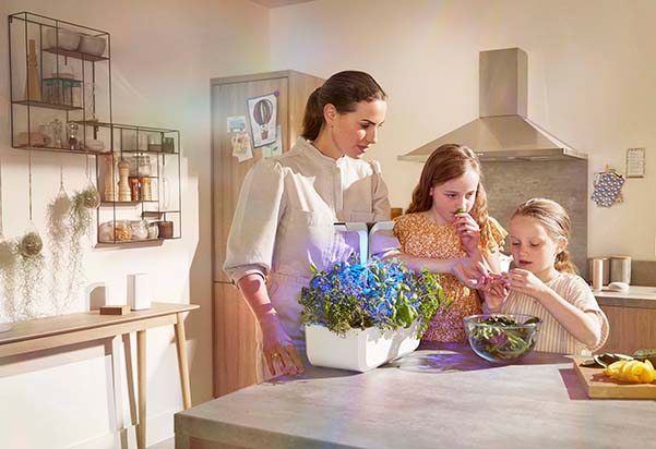 A mother and two children gathered around a Veritable smart indoor garden on a kitchen counter, with purple grow lights illuminating fresh herbs, in a bright modern home setting.
