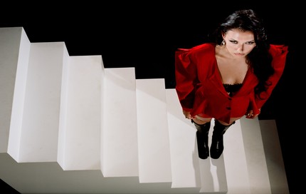 Nieve Ella in a red blazer and black knee-high boots standing on a white staircase, looking up at the camera in a promotional image for her tour