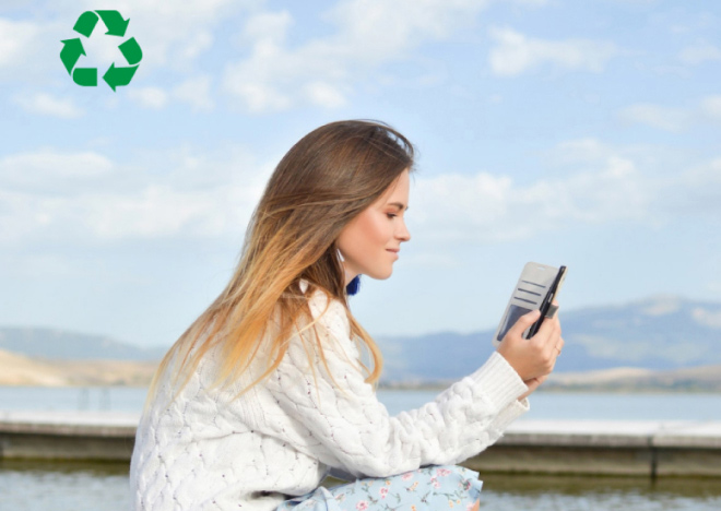 A young woman with long, light brown hair is looking at her phone outdoors beside a lake with mountains in the background under a blue sky. A green recycling symbol is in the top left corner of the image.