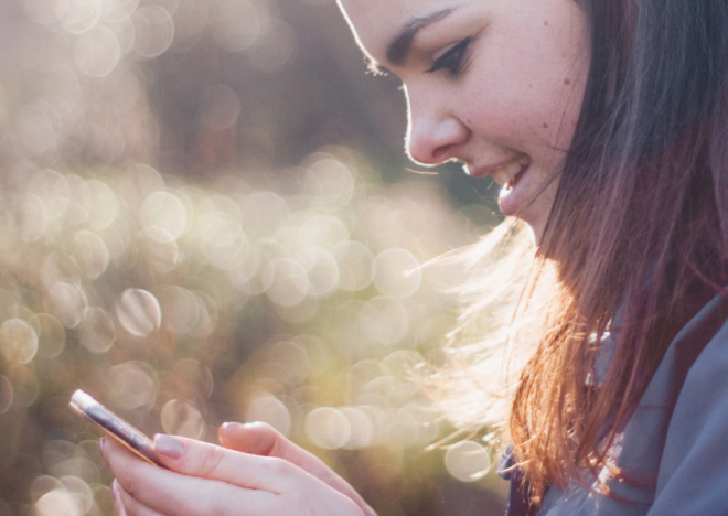 A smiling young woman with long hair is looking down at a smartphone she is holding with both hands. Her profile is visible, in an outdoor setting with sunlight.