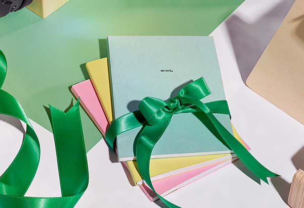 A stack of pastel-coloured notebooks tied together with a green satin ribbon bow, arranged on a light green surface with scattered ribbon, styled as a gift.