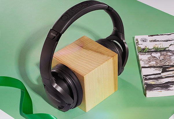 A pair of black Panasonic over-ear wireless headphones resting on a wooden block, displayed on a green surface with a green ribbon and printed photos nearby.