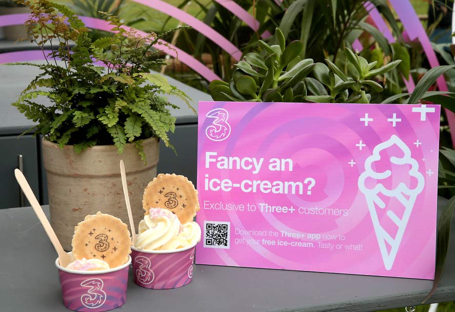 Two small cups of ice cream, each topped with a cookie, are sitting on a surface next to a pink sign. The sign has the Three logo and asks "Fancy an ice-cream?" It also features a QR code to avail of the offer exclusive to Three+ customers. Two small cups of ice cream, each topped with a cookie, are sitting on a surface next to a pink sign. The sign has the Three logo and asks "Fancy an ice-cream?" It also features a QR code to avail of the offer exclusive to Three+ customers.
