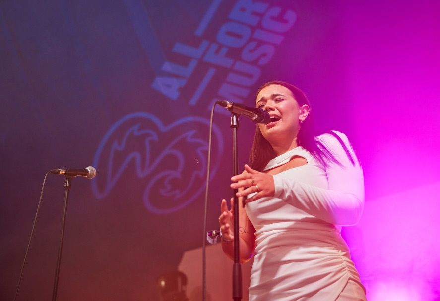 A female singer with long dark hair, performs on the Three Music stage. The stage is bathed in purple and pink lights, and a projection on the back wall reads "ALL FOR MUSIC" with the Three logo underneath. A female singer with long dark hair, performs on the Three Music stage. The stage is bathed in purple and pink lights, and a projection on the back wall reads "ALL FOR MUSIC" with the Three logo underneath.
