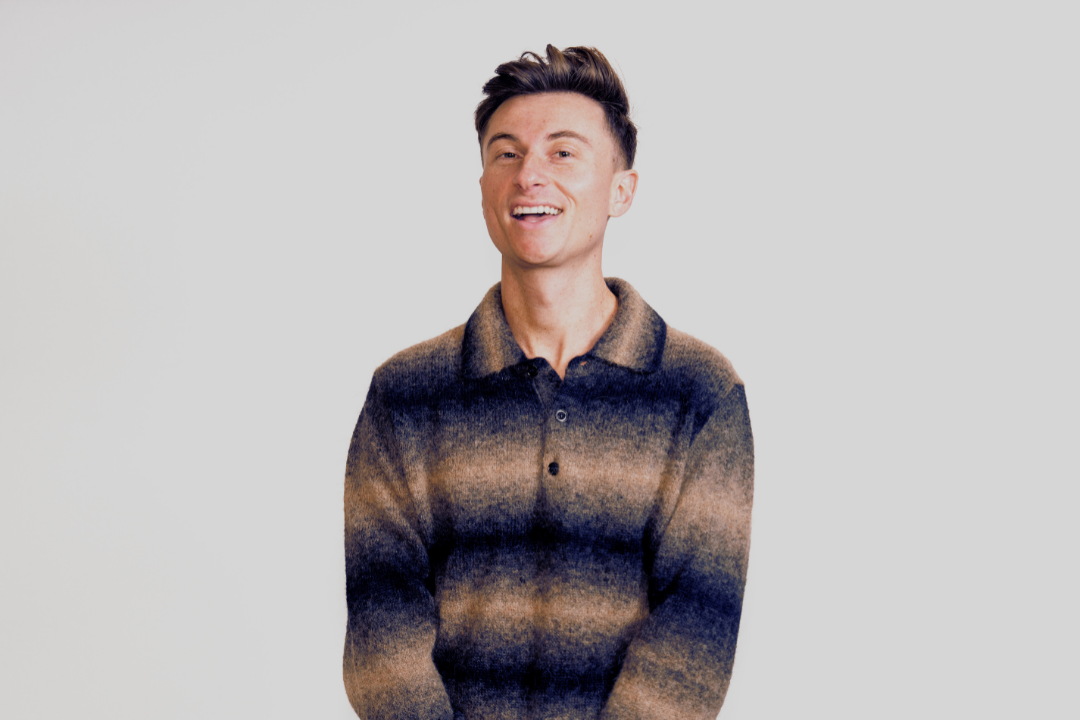 Trevor Wallace smiling in a striped knit sweater against a plain grey background in a promotional image for his The Alpha Beta Male tour.