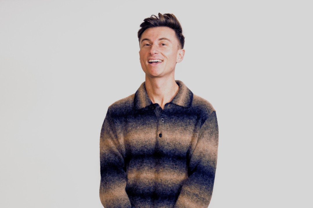 Trevor Wallace smiling in a striped knit sweater against a plain grey background in a promotional image for his The Alpha Beta Male tour.