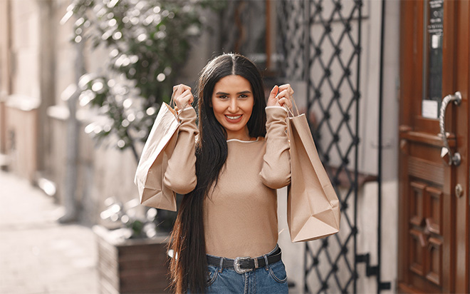 A smiling woman with long, dark hair stands outdoors, holding two light-colored shopping bags. A smiling woman with long, dark hair stands outdoors, holding two light-colored shopping bags.