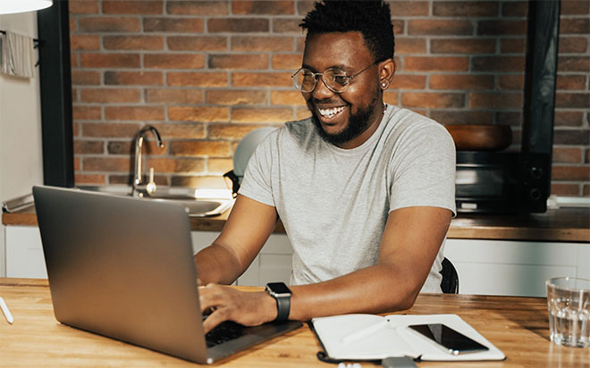 A man with a beard and glasses is seating at a wooden table, typing on a silver laptop. To his right, there is an open notebook, a smartphone, and a glass of water. In the background, a brick wall and kitchen fixtures are visible. A man with a beard and glasses is seating at a wooden table, typing on a silver laptop. To his right, there is an open notebook, a smartphone, and a glass of water. In the background, a brick wall and kitchen fixtures are visible.