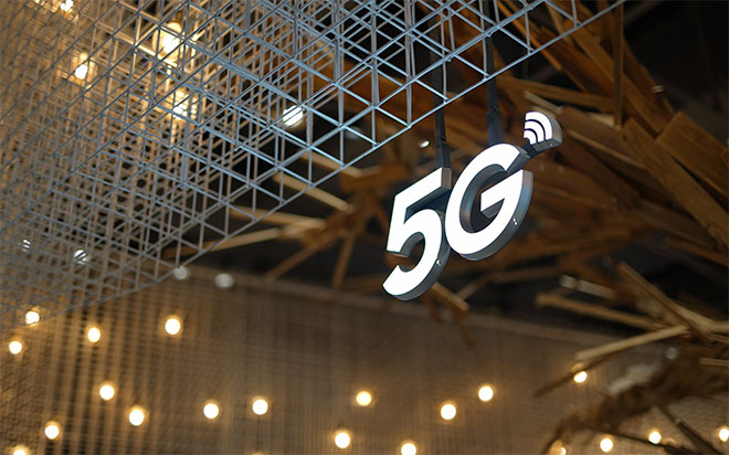 A glowing "5G" logo with a Wi-Fi signal icon is suspended, with a metal grid structure in the upper left foreground. Warm, blurred lights are visible in the background. A glowing "5G" logo with a Wi-Fi signal icon is suspended, with a metal grid structure in the upper left foreground. Warm, blurred lights are visible in the background.