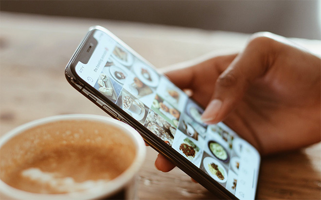 A hand holds a smartphone at an angle, with the screen displaying a grid of images. In the foreground, a white cup with coffee remnants is partially visible on a wooden table. A hand holds a smartphone at an angle, with the screen displaying a grid of images. In the foreground, a white cup with coffee remnants is partially visible on a wooden table.