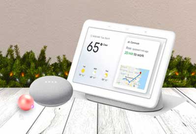 A Google Nest Hub displaying weather and commute information alongside a Google Nest Mini smart speaker, styled in a festive Christmas setting with pine garland and baubles.