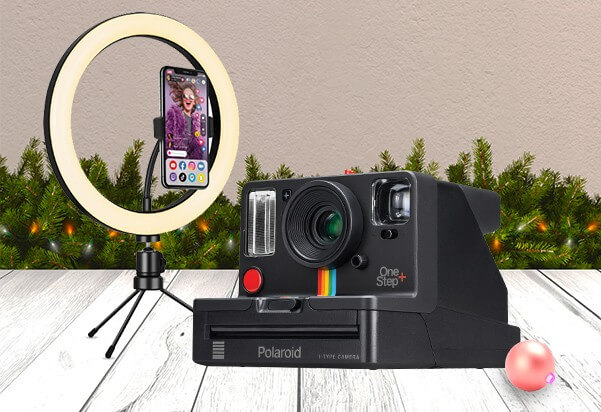 A Polaroid OneStep+ instant camera alongside a LED ring light with a smartphone mount on a mini tripod, displayed in a festive Christmas setting with pine garland and a bauble.