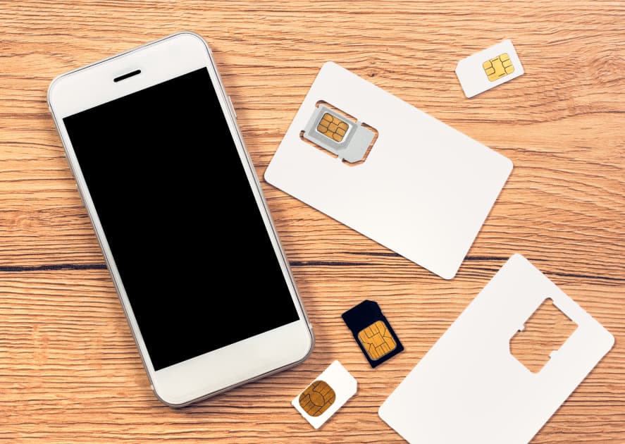 An overhead shot shows a white smartphone with a black screen resting on a wooden surface. Scattered around the phone are various white SIM card holders, some with SIM cards partially punched out, and several small SIM cards themselves. An overhead shot shows a white smartphone with a black screen resting on a wooden surface. Scattered around the phone are various white SIM card holders, some with SIM cards partially punched out, and several small SIM cards themselves.
