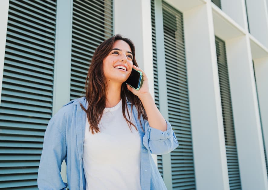 A smiling young woman with long, dark hair is talking on a smartphone, holding it to her ear. The background features a modern building with horizontal white slats and vertical pillars. A smiling young woman with long, dark hair is talking on a smartphone, holding it to her ear. The background features a modern building with horizontal white slats and vertical pillars.
