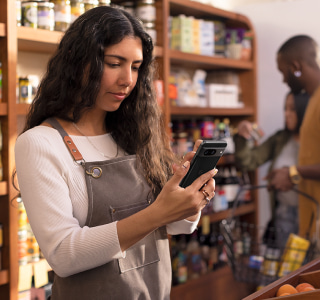 A person with long, dark, wavy hair wears an apron and looks at a smartphone in a shop setting. A person with long, dark, wavy hair wears an apron and looks at a smartphone in a shop setting.