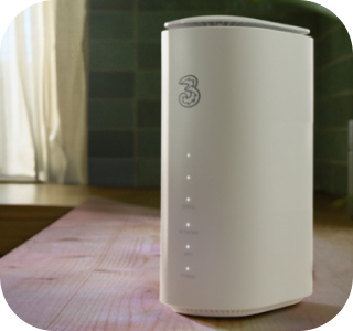 A Three router with a "3" logo and five indicator lights on its front is positioned on a light wooden table. In the blurred background, a window and a tiled wall are visible. A Three router with a "3" logo and five indicator lights on its front is positioned on a light wooden table. In the blurred background, a window and a tiled wall are visible.