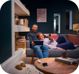 A smiling couple relaxes on a gray sofa in a living room. The man sits upright, holding a tablet, while the woman reclines with her head on his shoulder, taking a selfie with her phone. A smiling couple relaxes on a gray sofa in a living room. The man sits upright, holding a tablet, while the woman reclines with her head on his shoulder, taking a selfie with her phone.