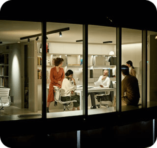 View from outside at night of an office, a number of people are gathered in a well lit room having a conversation. View from outside at night of an office, a number of people are gathered in a well lit room having a conversation.