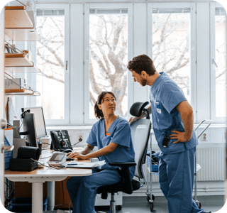 Two people wearing scrubs converse at a desk, sharing information in a healthcare setting. Two people wearing scrubs converse at a desk, sharing information in a healthcare setting.