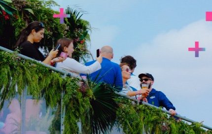 A group of people on a brightly coloured walkway or bridge, with lush green foliage. People are standing, some looking at phones, others interacting. The background is a soft blue with scattered white and pink cross shapes.