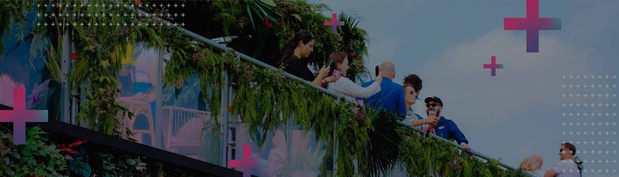 A group of people on a brightly coloured walkway or bridge, with lush green foliage. People are standing, some looking at phones, others interacting. The background is a soft blue with scattered white and pink cross shapes.