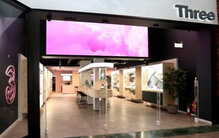 The entrance of a "Three" store features a large digital display with pink clouds, a prominent neon "3" logo on the left wall, and an interior with light wood and dark accents, showcasing electronic devices.