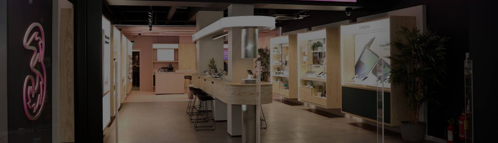 A sleek, modern Three store interior with a light wood and dark grey colour scheme. On the left a neon "3" logo glows pink. Display cases with electronic devices and accessories line the walls and a light-coloured counter with stools extends into the room