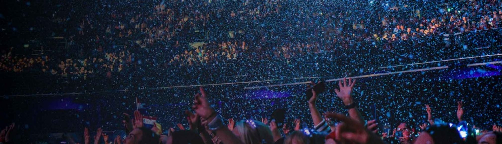 A massive, energetic crowd at a live concert with hands raised in the air amidst a heavy shower of blue confetti.
