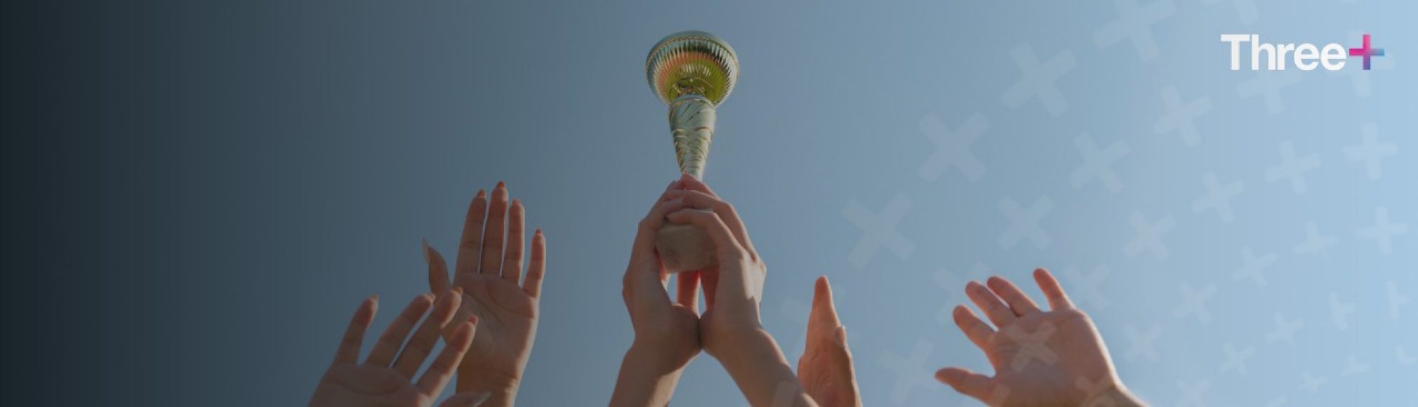 Multiple hands raised in celebration against a blue sky, with the center hands holding up a golden trophy. The "Three+" logo is in the top right corner.