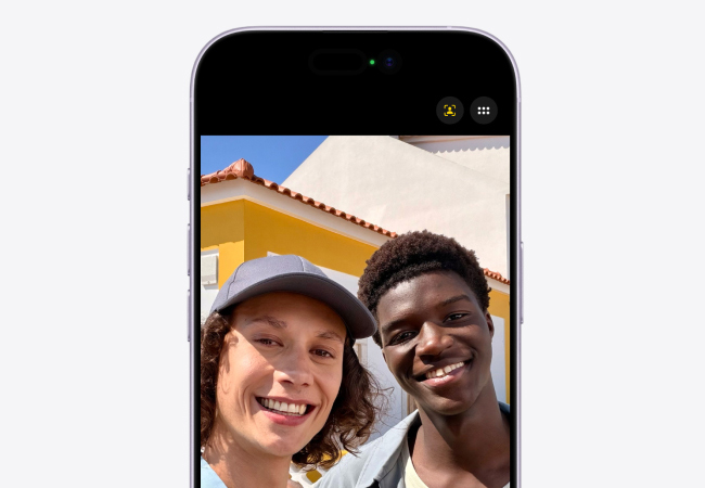 A selfie on an iPhone 17 screen showing two young people smiling. The background is a yellow and white building.