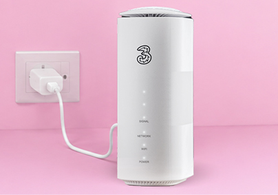 A white Three mobile broadband router plugged into a wall outlet, set against a solid pink background.
